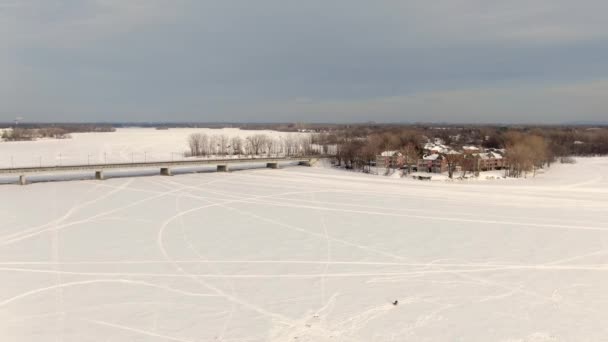 セント エスタシュとラバル オウエストと2つの側面を結ぶ橋の間の凍った川をゆっくりと回転するドローンビデオ 人々は氷釣り — ストック動画