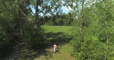 Havada drone görüntüsü, ağaçlar arasında yürüyen bir bayanı takip ediyor, bir parkın içinde, yaz boyunca.