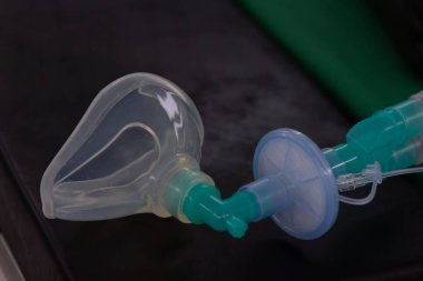 Klinik ortamda karanlık bir yüzeye yerleştirilmiş yeşil bir solunum tüpüne bağlı temiz bir tıbbi oksijen maskesinin yakın çekimi.
