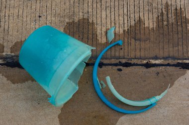 Kırıldıktan sonra etrafa saçılmış ıslak beton yüzeyde parçalanmış mavi plastik bir kova.