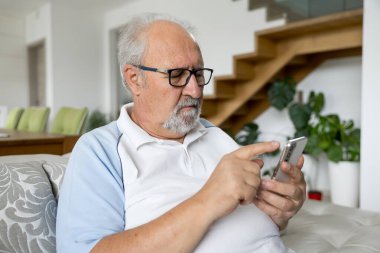 Gözlüklü yaşlı bir adam kanepeye oturmuş, akıllı telefonunu parlak, modern bir oturma odasında kullanıyor..