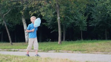 Küçük çocuk ellerini tutuyor ve çubukta mavi pamuk şeker yiyor. Panayır günü bir çocuk parkın yolunda yürüyor..