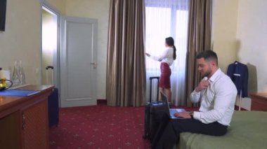 Beyaz gömlekli bir iş adamı otel odasında bir yatakta oturuyor. Tatilde bir dizüstü bilgisayarda uzaktan çalışma. Pencerenin yanında genç bir kız. Seyahat için bavul.