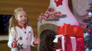 Gülümseyen sevimli küçük kız, kırmızı kutularla yeni yıl hediyeleriyle oynuyor. Noel süsleri ve şöminesi olan bir oda..