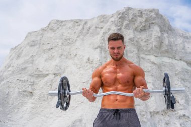 Güçlü bir vücut geliştirici. Elinde halterle dağların arkasında.