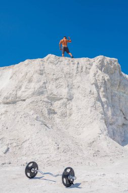 Atletik erkek vücut geliştirici beyaz bir dağda mavi gökyüzüne karşı poz veriyor. Önplanda metal çubuk
