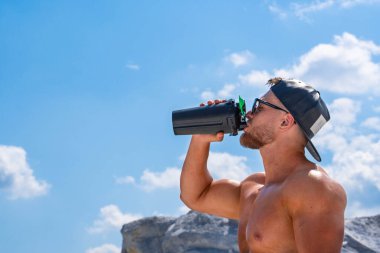 Beyzbol şapkalı erkek sporcu, bulutlu mavi gökyüzüne karşı şişeden enerji içeceği içiyor.