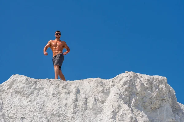 Çıplak vücutlu bir erkek vücut geliştirici beyaz bir dağda kaslarını gösteriyor. Arka plandaki mavi gökyüzü