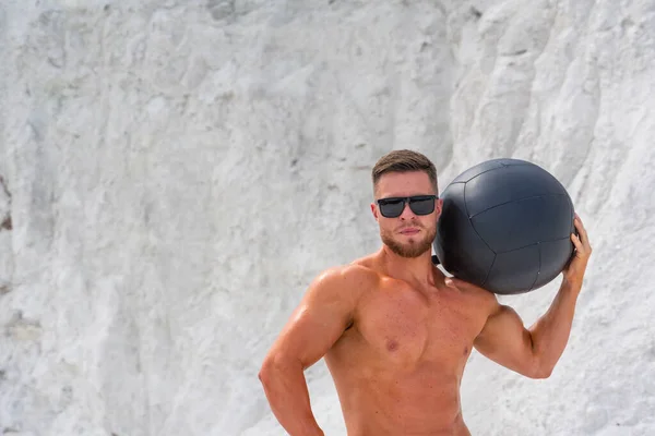 Gözlüklü ve kaslı bir adam omzunda siyah bir fitness topu tutuyor. Arka planda beyaz dağ var. Doğada fitness dersi