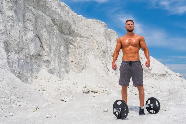 stock image Inflated strong athletic build man with a naked torso in shorts near the barbell against a background of white mountains and blue sky