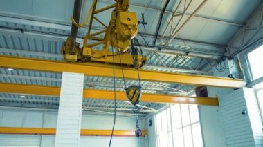Rehber korkuluklar boyunca bir elektrikli vinç hareket ediyor tavanın altından fabrika odasına doğru. Büyük sarı vinç kancası sallanıyor.