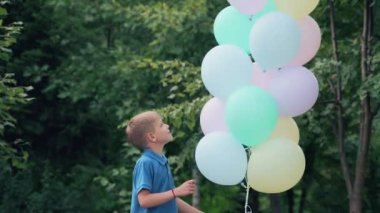 Mavi tişörtlü mutlu çocuk, yaz parkında renkli helyum balonlarıyla oynuyor. Çocuk atlayışı.