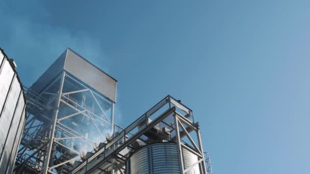 グレインエレベーターの金属構造 明確な青い空に対して穀物乾燥機のパイプからの煙 作物の貯蔵のための特別な穀物のサイロ — ストック動画