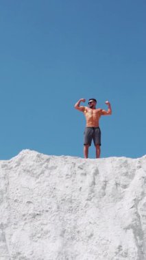 Gözlüklü güçlü bir adam kaslı vücudunu gösterir. Mavi gökyüzüne karşı beyaz bir dağda vücut geliştirme. Dikey video.