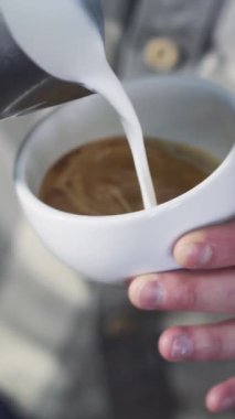 Profesyonel barista beyaz bardağa süt köpüğü dökerek mükemmel bir kapuçino yaratıyor ve güzel bir desen oluşturuyor. Dikey video.