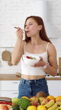 Mutfaktaki mutlu kız tabaktan taze sebze salatası yiyor. Vejetaryen yemekleri için sebze ve meyvelerle dolu bir masa. Dikey video.