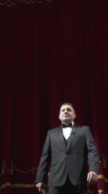 Takım elbiseli, papyonlu ve elinde mikrofonlu bir şovmen tiyatronun sahnesinde gösteri yapıyor. Dikey video.