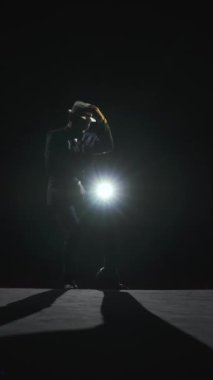 Takım elbiseli yetişkin bir adam sahne ışıkları altında bir tiyatronun karanlık sahnesinde dans eder. Şapkalı bir dansçı silueti. Dikey video.