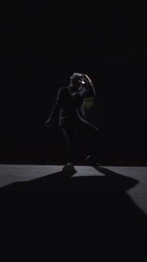 Tiyatroda dans eden takım elbiseli ve şapkalı yetişkin bir adamla büyük bir karanlık sahne. Soffit light. Steadicam çekimi.
