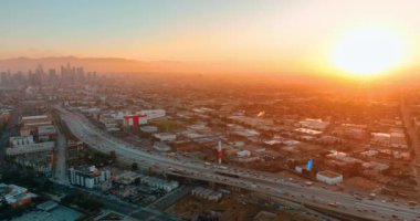 Gün batımında Los Angeles 'ta devasa bir otoyol kavşağının havadan panoramik görüntüsü. Şehir merkezinde gökdelenler ve ufukta görünen tepeler.