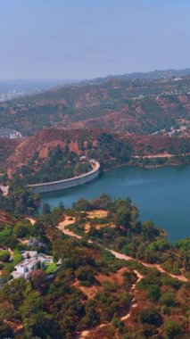Hollywood Reservoir barajının su dolu havadan görünüşü. Tepelerdeki konut binaları ve bitki örtüsü. Dikey video.