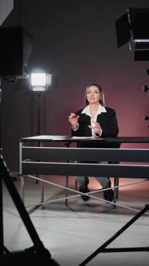 Televizyon stüdyosunun sahne arkasında talk show çekimleri. Stüdyoda kameraya konuşan genç bir TV sunucusu. Kameralı ve ışıklandırmalı stüdyo tripodları. Dikey video.