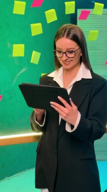 Siyah ceketli ve gözlüklü bir kız etiketli bir tahtanın yanında duruyor ve elinde bir tablet tutuyor. Gülümseyen bir kadının portresi. Dikey video.