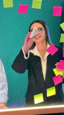 Ofisteki iş kadını renkli yapıştırmalarla şeffaf bir cam tahtayı siliyor. İşteki kız gülümsüyor. Dikey video.