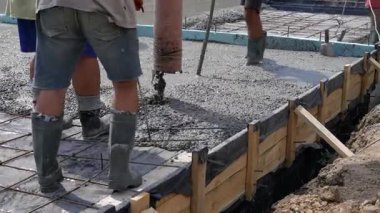 Kauçuk çizmeli işçiler güçlendirilmiş demir ızgaralı bir evin temelleri için beton dökerler..