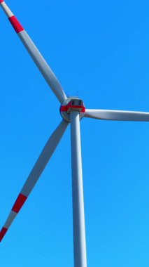 Rüzgâr türbinine yakın çekim. Bıçaklar dönüyor ve elektrik üretiyor. Mavi gökyüzü arkaplanına karşı jeneratörün arka görüntüsü. Dikey video.