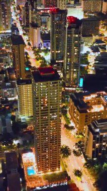 Oahu adasında Honolulu 'nun ışıldayan gece sokakları. Waikiki 'de ışıkları olan uzun otel binaları. Arabalar yolda. Hava görüntüsü. Dikey video.