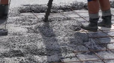 Beton, lastik çizme giyen iki işçi tarafından çelik inşaat demiri ızgarasının üzerine dökülüyor. Yüzey ıslak ve şebeke yapısal destek sağlıyor..