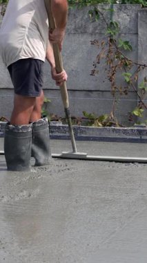 Lastik çizmeli bir insan, uzun saplı bir vida kullanarak yeni dökülmüş betonu düzleştiriyor. Yüzey ıslak ve arka planda bitki örtülü beton bir duvar var. Dikey video.