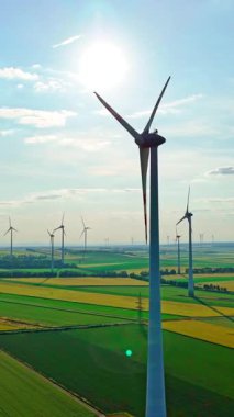 Ön planda uzun bir rüzgâr türbini ve pervanelerin arkasında da güneş parlaması. Tarım alanlarının altında yeşil ve sarı tonlarda çeşitlilik var. Hava görüntüsü. Dikey video.