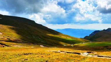 Parçalı bulutlu bir gökyüzünün altında Romen Karpatlar 'ında yuvarlanan tepeler ve çimenli vadiler. Dolambaçlı bir patika araziyi kesiyor, uzak mavi tepelere doğru gidiyor..