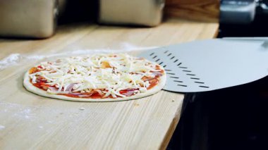 Çiğ pizza hamuru ve ahşap mutfak masasına peynir serpiştirilmiş çeşitli malzemeler. Şef pizzayı fırın spatulasıyla alıyor. Yakın plan..