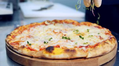 Şef mikroyeşilliklerle süsleyip süslüyor. İçi malzemelerle dolu taze bir pizza. Mutfaktaki masanın üzerinde ahşap bir tahta üzerinde yuvarlak pişmiş pizza..