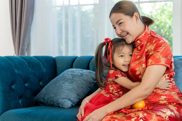 Happy Asian family of mother and little daughter wearing traditional costumes sitting on couch at home,  give auspicious orange to mother, Hug and smile together on Chinese New Year day