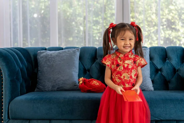 Stock image Portrait Happy Asian little girl wearing chinese traditional costumes sitting on couch with red envelopes in hands on Chinese New Year day, copy space