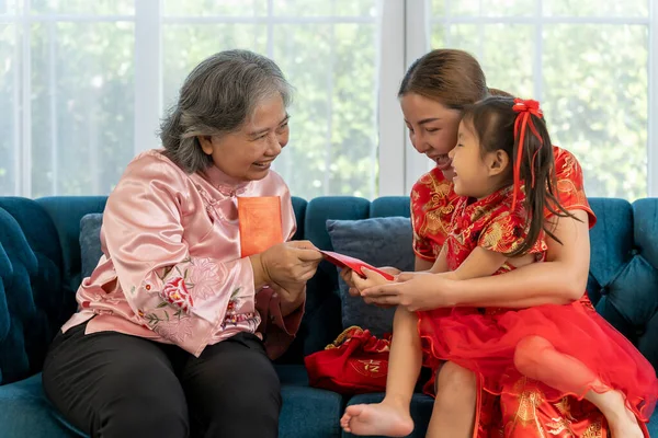 Happy Elderly woman sitting on sofa with daughter and granddaughter take of a banknote from red envelope and happy to show, They laugh with fun together on Chinese New Year
