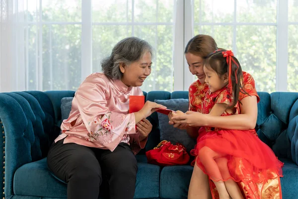 Happy Elderly woman sitting on sofa with daughter and granddaughter take of a banknote from red envelope and happy to show, They laugh with fun together on Chinese New Year
