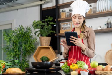 Aşçı şapkası ve önlüğü takan Asyalı güzel bir kadın yemek pişirme, elektrikli soba, yumurta, sebze ve mutfak gereçleri reyonunda yemek tarifleri izlemek için tablet kullanıyor.