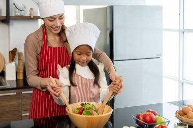 Güzel anne ve küçük kızından oluşan neşeli bir aile. Gülümsüyor ve evde sağlıklı yemekler pişirirken birlikte salata sebzeleri yapmanın tadını çıkarıyor.