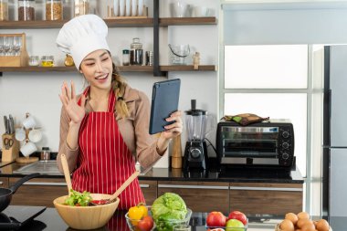 Aşçı şapkası ve önlüğü takmış güzel Asyalı kadın portresi mutfakta yemek pişirirken dijital tablet görüntüsü kullanın. Tencere ve elektrikli soba, yumurta ve teçhizat tezgahta. Boşluğu kopyala