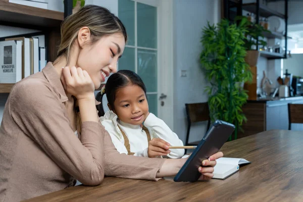 Beautiful mother teaching her lovely daughter to do homework with digital tablet in living room at house, they talking and smile with happy together, copy space