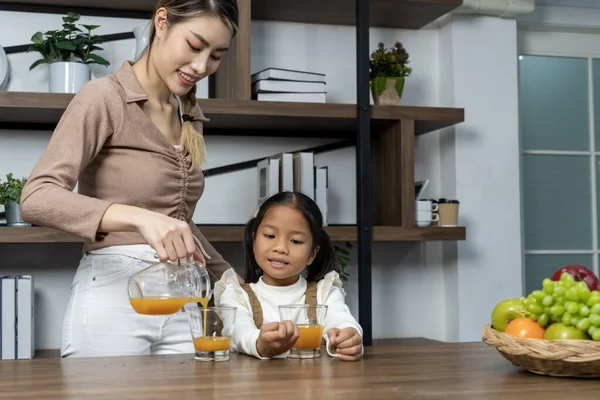 stock image Beautiful mother pouring fresh orange juice into glass for her lovely daughter on table in kitchen at house, they talking and smile with happy together, copy space