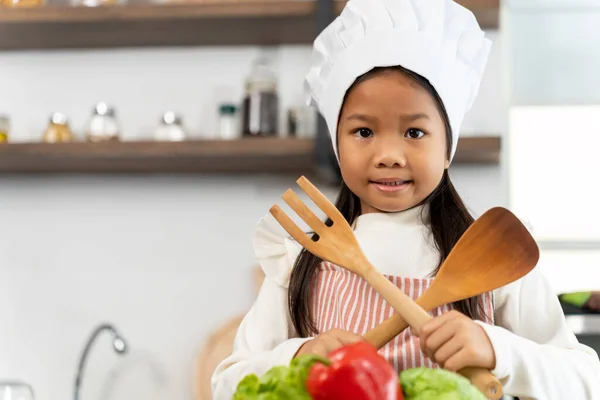 用双手拿着木勺和叉子的可爱女孩的画像 微笑着看着相机 厨房柜台上的蔬菜 鸡蛋和炊具 复制空间 图库图片