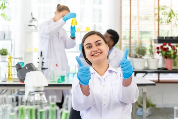 研究小组的同事们在她身后的实验室里成功地完成了一个年轻科学家或研究人员带着医用手套竖起大拇指的画像 — 图库照片