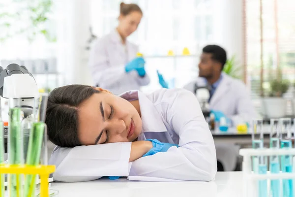 中国日报 的报道 在实验室里 当同事在她身后的模糊背景下工作时 年轻的女科学家在桌子上昏昏沉沉地睡着了 — 图库照片