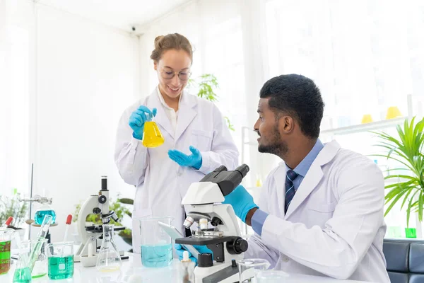 stock image Portrait of a young scientist or researcher with medical gloves showing samples results of her experiment in lab with successful to her colleague male in laboratory, microscope and scientific equipment place on desk